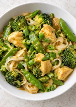 Coconut curry chicken with broccoli, snap peas, scallions, and a small portion of rice noodles in a white bowl