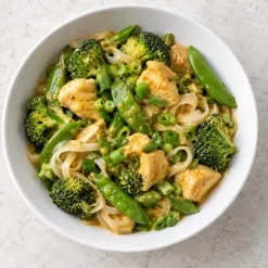 Coconut curry chicken with broccoli, snap peas, scallions, and a small portion of rice noodles in a white bowl