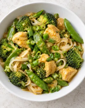 Coconut curry chicken with broccoli, snap peas, scallions, and a small portion of rice noodles in a white bowl